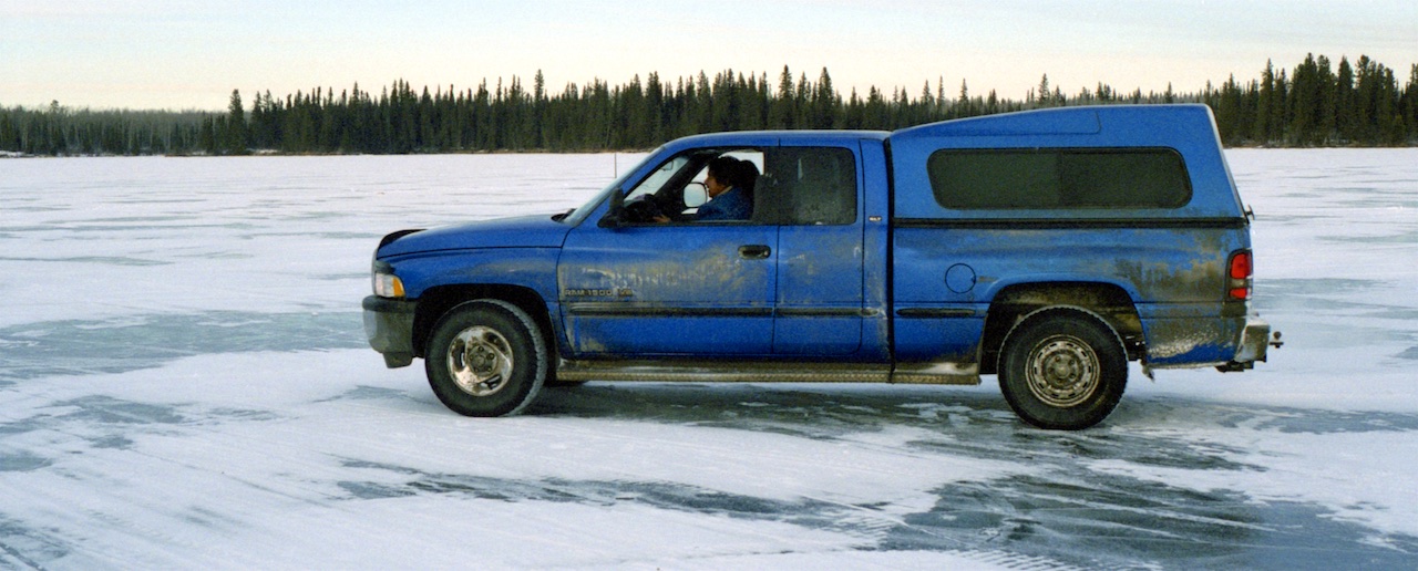 A dirty blue pickup truck with occupants parked on a frozen lake.